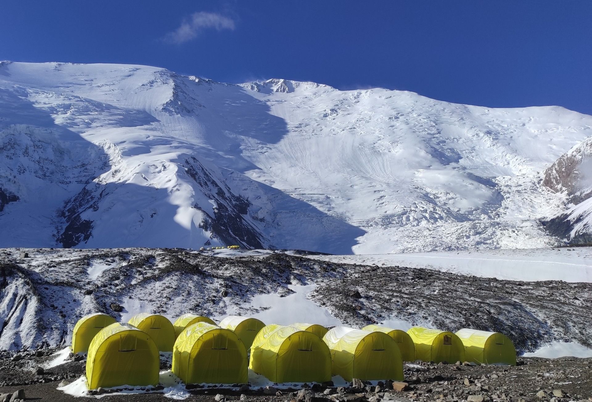 Un massiccio innevato sotto un cielo blu intenso, con le tende gialle di un campo ai suoi piedi.