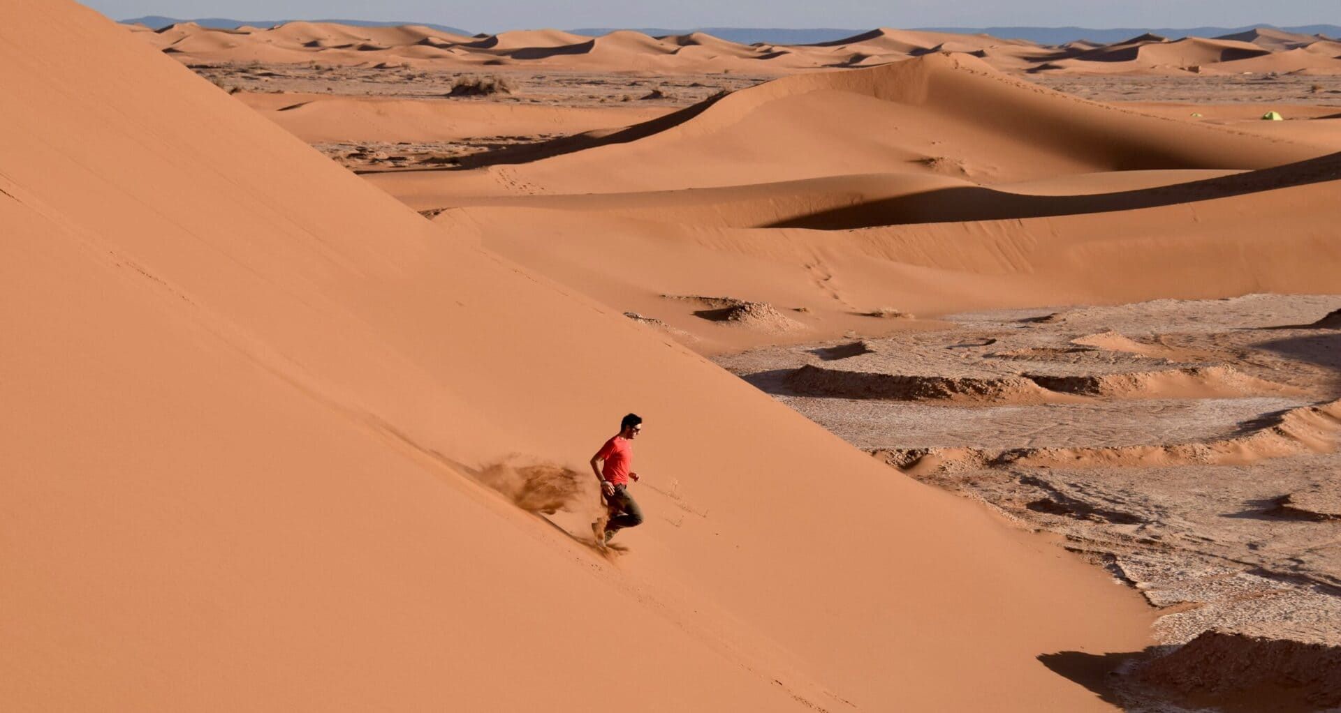Una persona corre giù da una grande duna arancione, il deserto fino all’orizzonte.